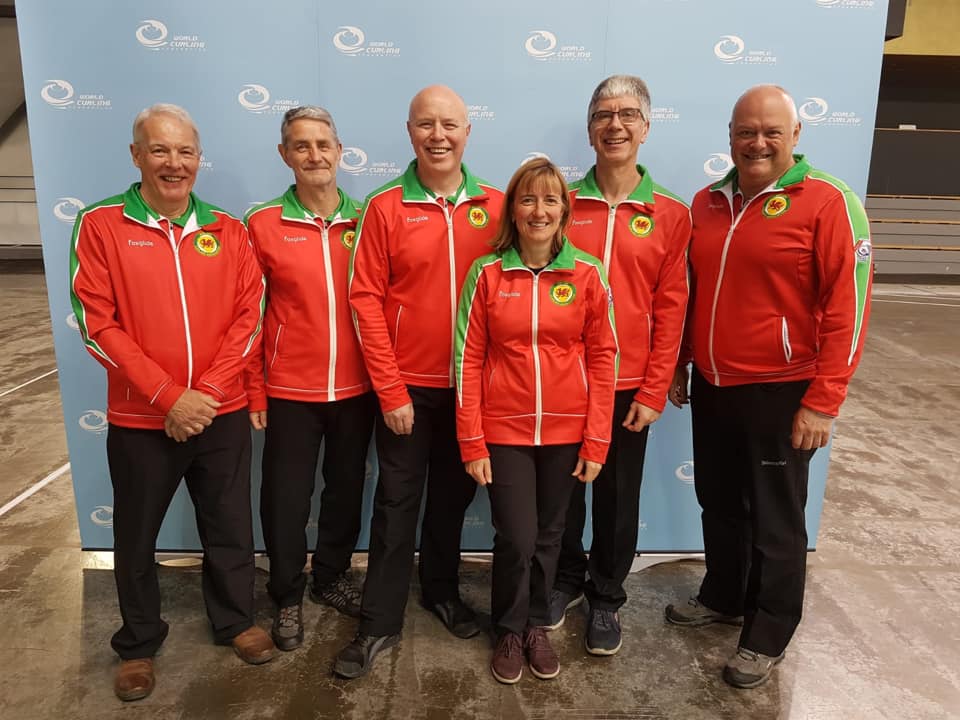 Five men and one woman stand in front of a blue background, all wearing black trousers with red and green jackets