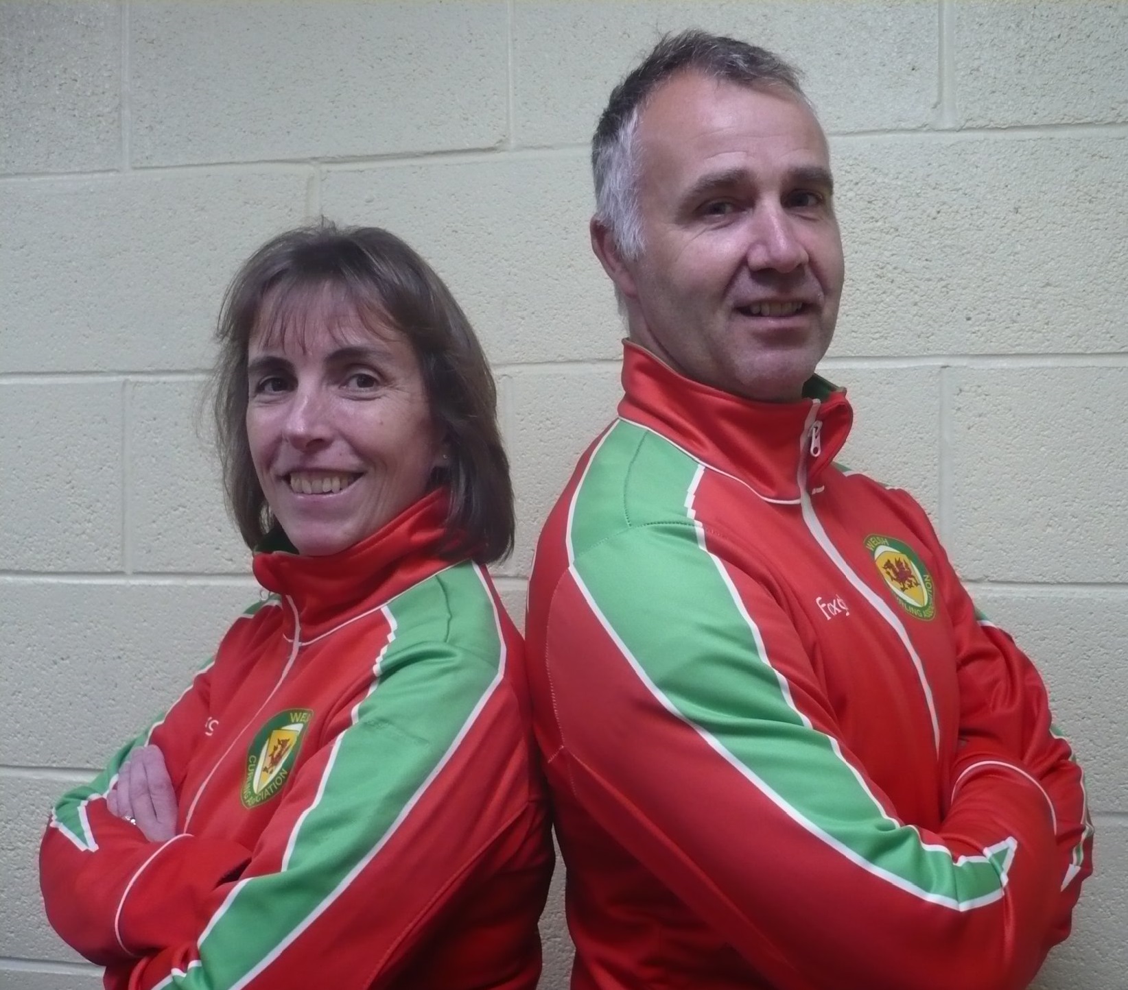 Photograph of a man and a woman wearing red and green jackets in front of a white brick wall